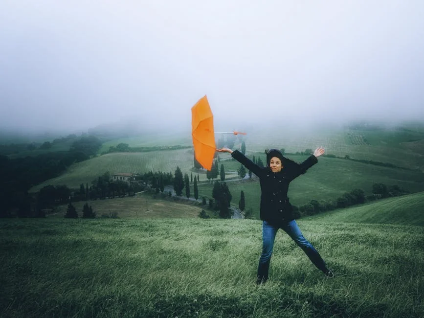 A woman holds an orange umbrella in a foggy countryside, showcasing nature and weather dynamics. - best compact umbrellas for windy weather