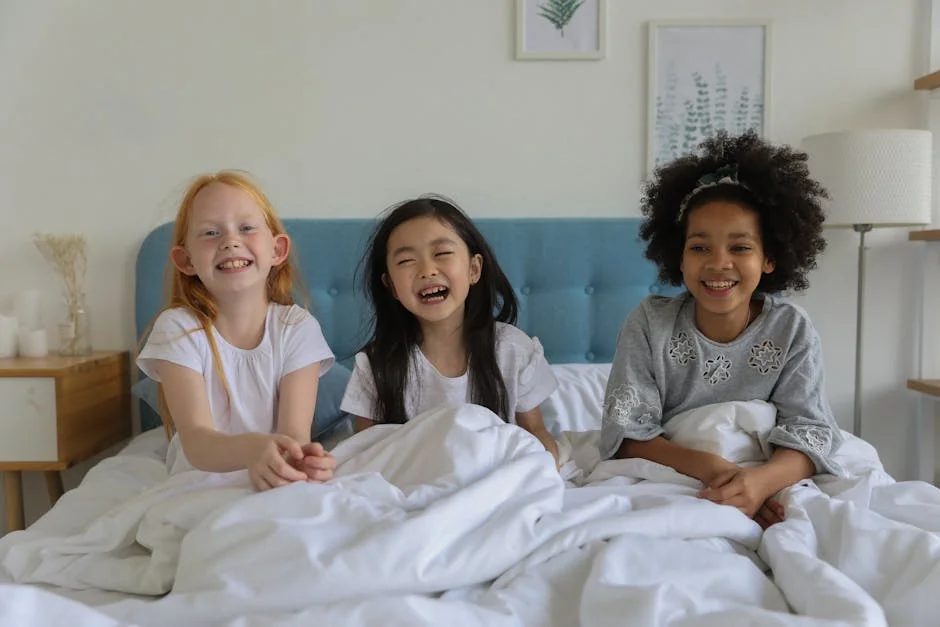 Laughing multiethnic best friends enjoying time during sleepover lounging under blanket in bed - best waterproof mattress protectors for kids