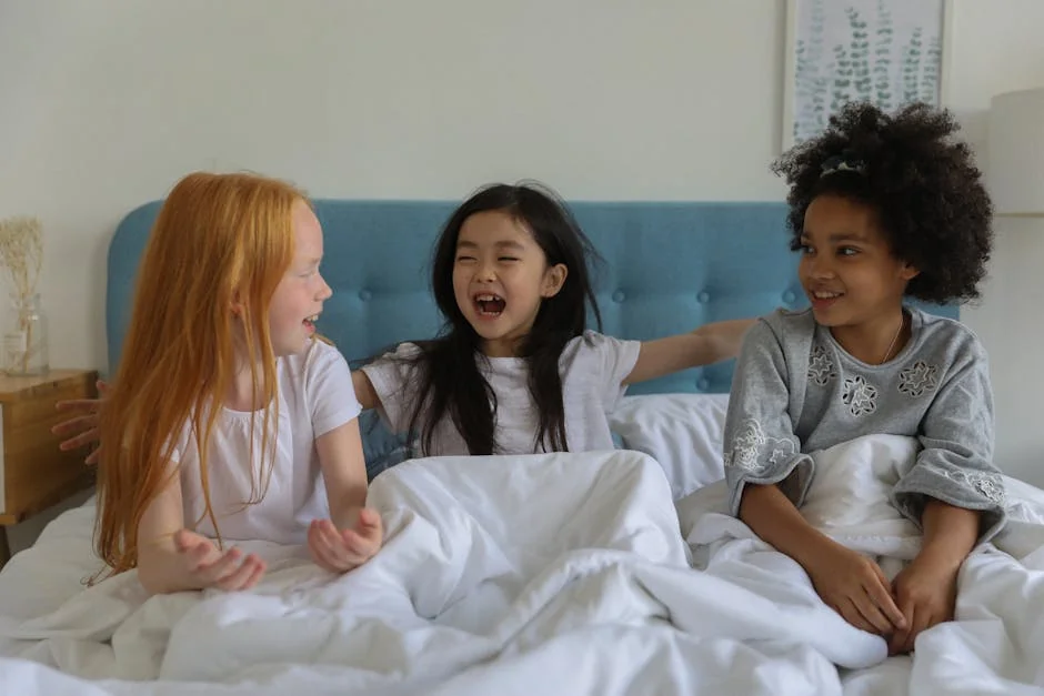 Adorable diverse girls laughing and chatting while sitting on bed under blanket at home - best waterproof mattress protectors for kids