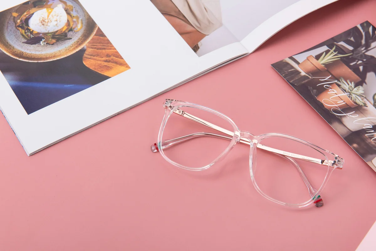 The glasses sport a clear frame adorned with a red floral accent, perfectly juxtaposed against a soft pink backdrop. The transparent frames harmonize with the magazine's warm hues, enhancing the contrast and visual appeal. - most stylish blue light blocking glasses for work