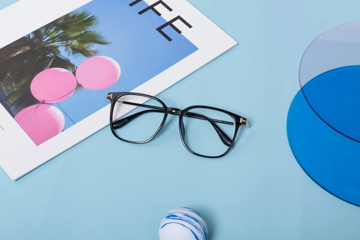 One unique pairs of eyeglasses are artfully arranged on a soft blue background. Accompanied by a magazine and a delicate flower, this display highlights the blend of creativity and refinement in modern eyewear. - most stylish blue light blocking glasses for work