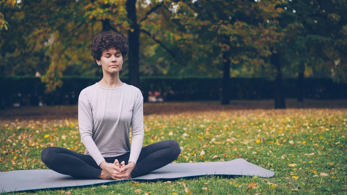 Flexible young lady is sitting on mat in yoga pose enjoying fresh air, peace and relaxation. Healthy lifestyle for urban people, active youth and autumn nature concept. - meditation wellness relaxation - top rated yoga mats for high intensity workouts